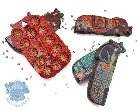 A collection of foldable wooden mancala boards with handcrafted batik patterns, along with cowrie shells used as game seeds. The boards display a variety of colors and patterns.