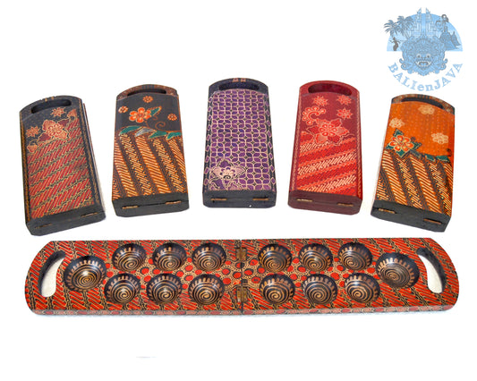 A collection of foldable wooden mancala boards with handcrafted batik patterns in various colors, along with cowrie shells and a batik canvas dust bag.