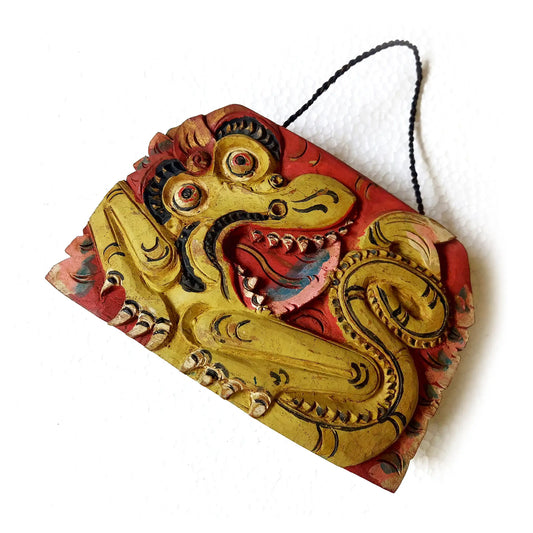 A hand-carved wooden bell depicting a mythological dragon with an open mouth, painted in yellow, red, and black tones, suspended by a black cord against a plain white background.