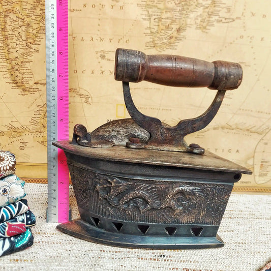 A vintage charcoal iron with a dark brown wooden handle, displayed next to a pink and silver ruler for scale against a vintage map background.
