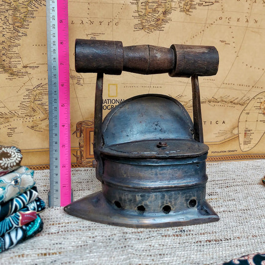 A vintage dark metal charcoal iron with a wooden handle, shown next to a pink and silver ruler for scale, set against a background of an old map and burlap fabric.