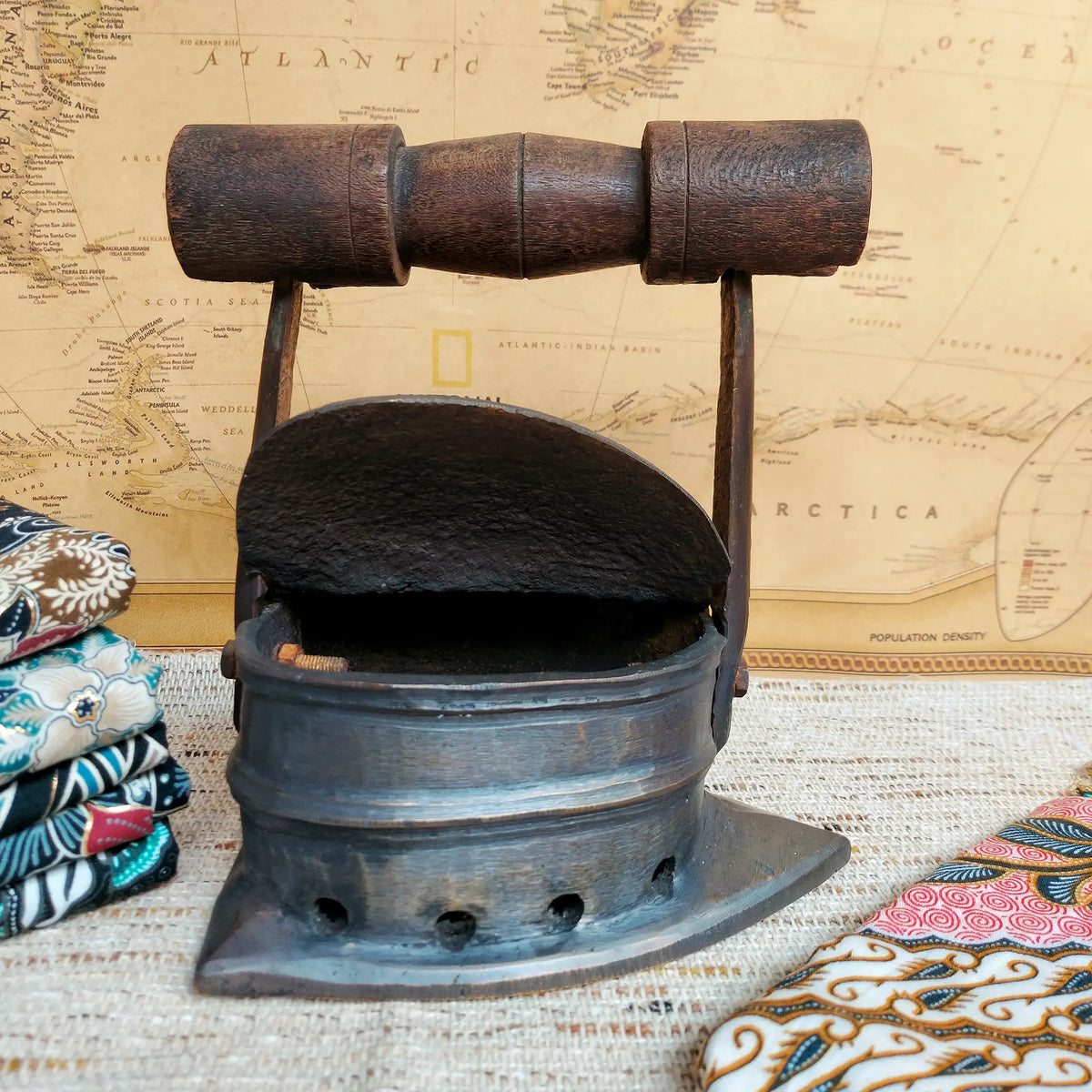 A vintage charcoal box iron with a dark brown wooden handle, displayed on a textured burlap surface with a vintage map and patterned textiles in the background.