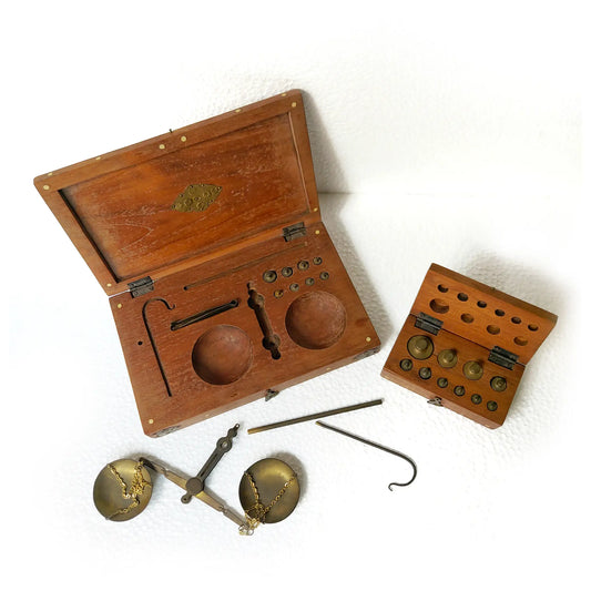 Vintage brass weight set with balance scales, hooks, and adjustable components, displayed with their wooden storage boxes on a white background.
