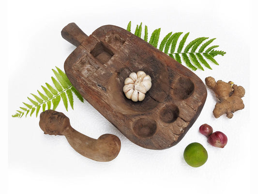 A rustic dark brown wooden mortar and pestle set arranged on a white background, styled with green fern leaves, onions, ginger, lime and red onions.