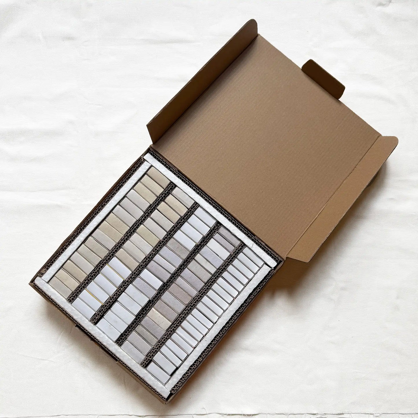 An open brown cardboard box containing multiple small rectangular matchboxes arranged in rows, photographed from above on a white fabric background.