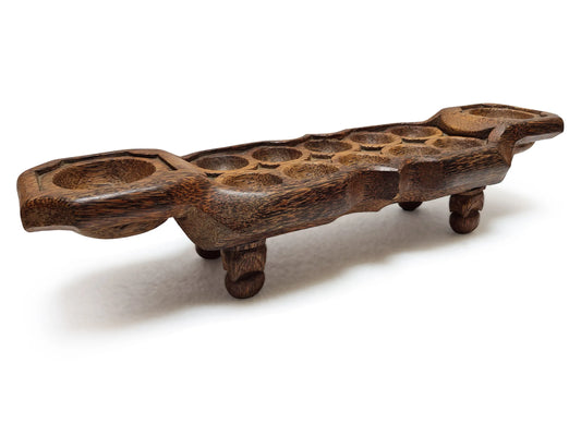 A dark brown carved wooden mancala game board multiple circular holes and four small legs, photographed on a white background.