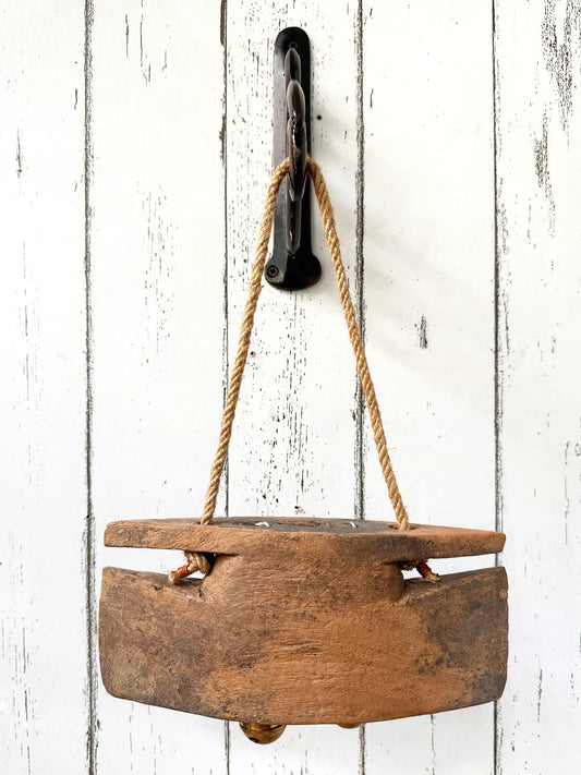 A old wooden bell, suspended by a jute rope from a black metal hook mounted on a white distressed wood plank wall.