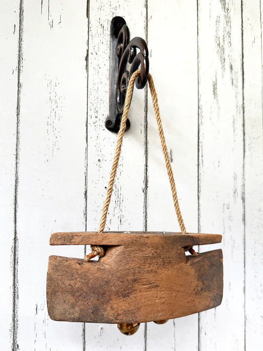 A rustic wooden bell with a natural jute rope handle hanging on a black metal wall hook, shown against a distressed white wood plank background.