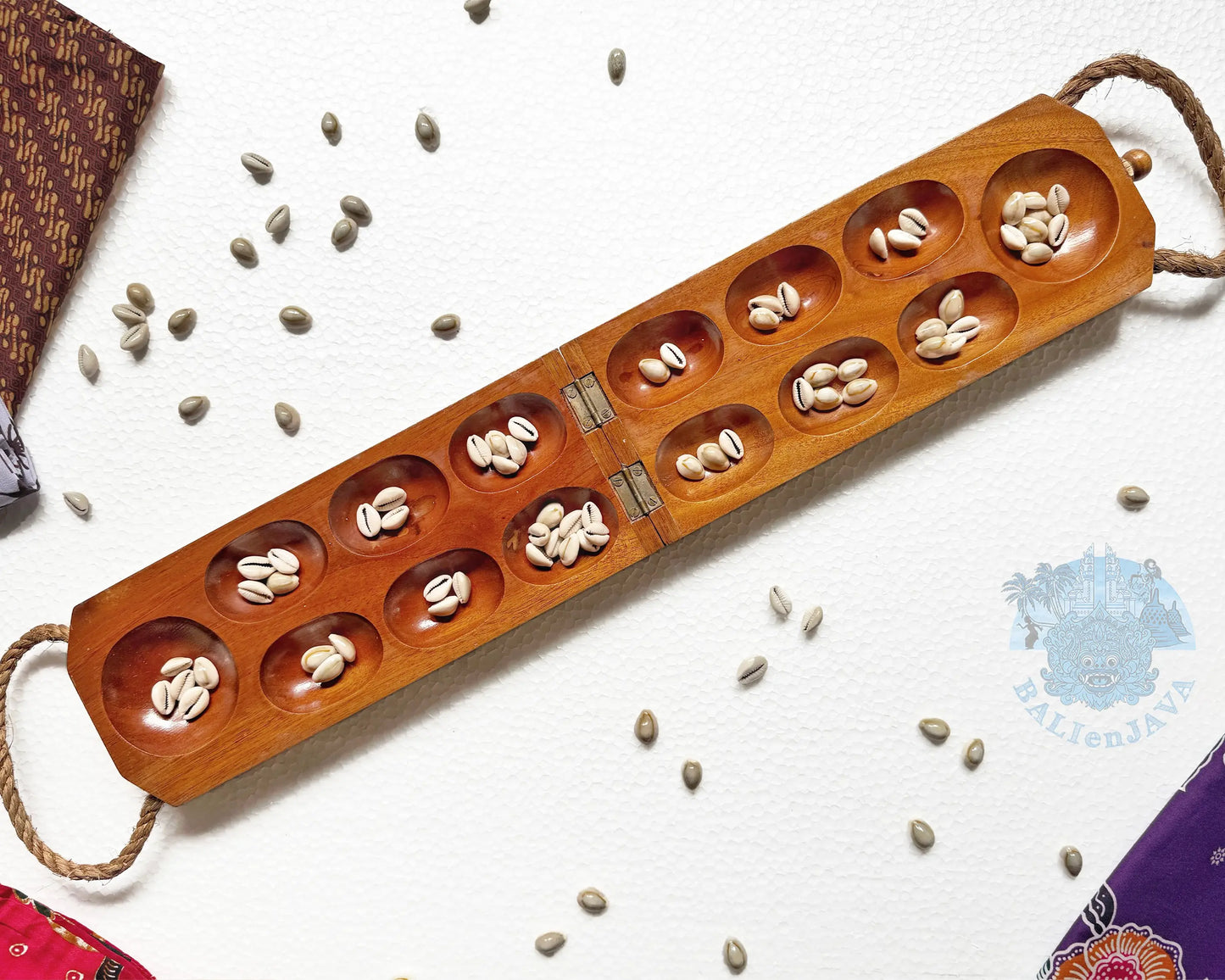 A wooden mancala game board with multiple oval compartments containing small white beans, photographed on a white textured background with scattered cowrie shells and small batik fabric pieces at the corners.