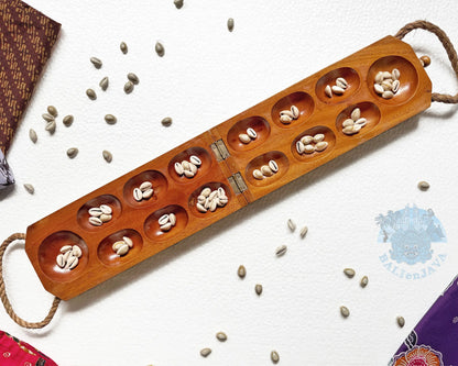 A wooden mancala game board with multiple oval compartments containing small white beans, photographed on a white textured background with scattered cowrie shells and small batik fabric pieces at the corners.