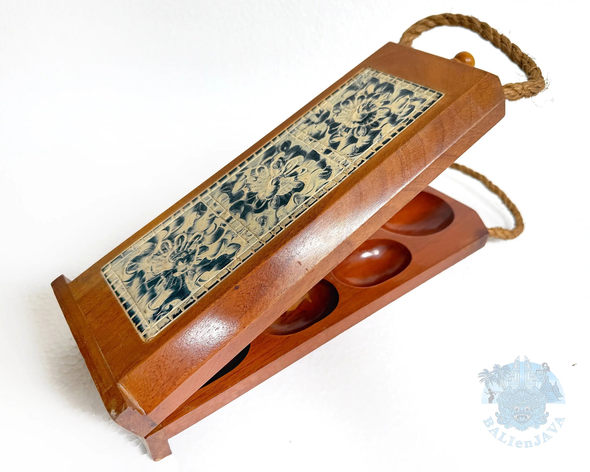 A wooden mancala board game with a hinged lid, featuring a decorative blue and beige floral pattern on the front panel and oval compartments visible inside. A rope handle is attached at the top, photographed on a plain white background.