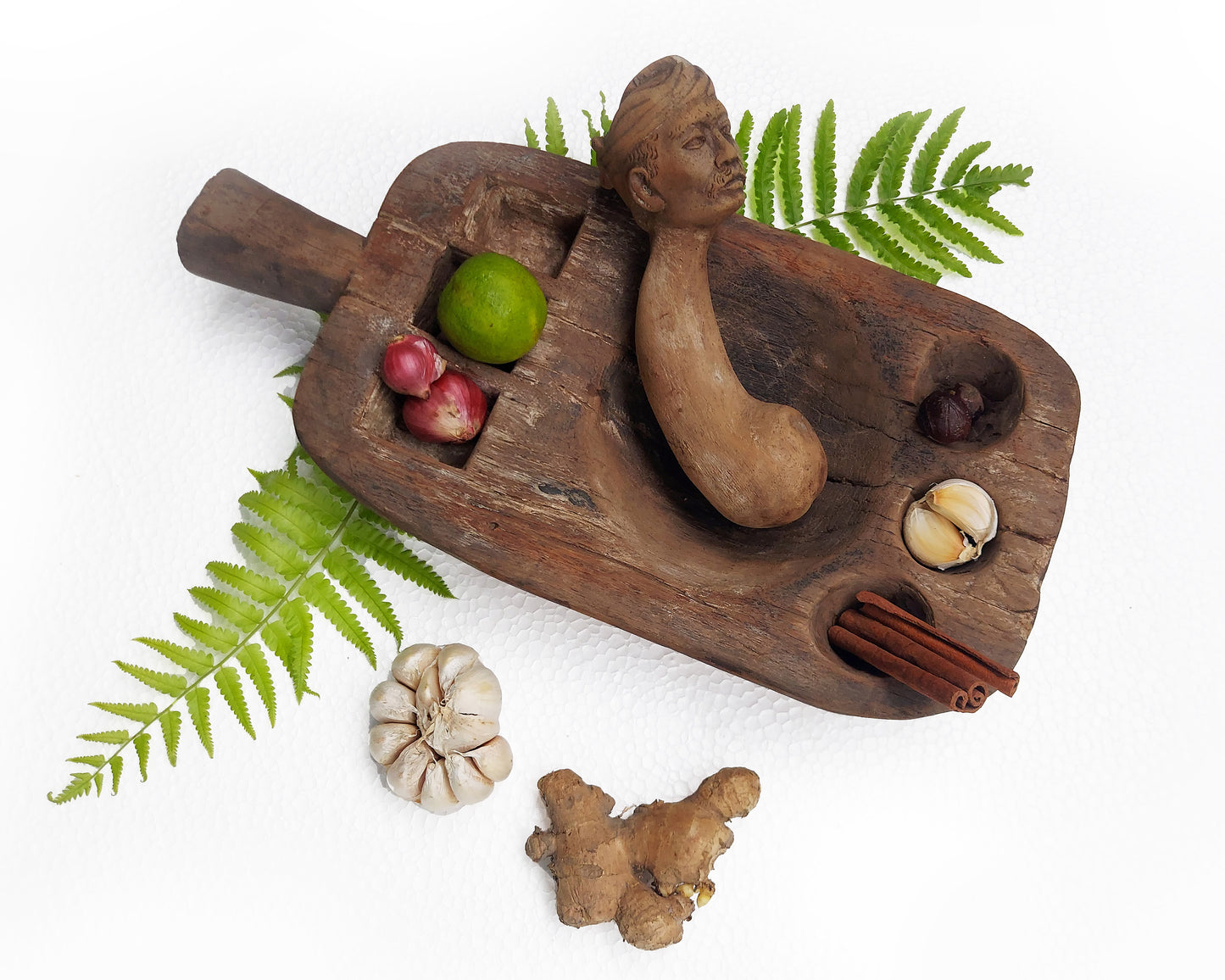 An old, hand-carved wooden mortar and pestle set with various spices and a traditional figure head on a white background.