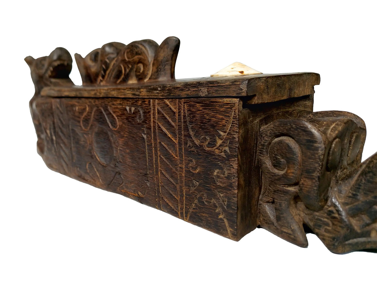 An antique carved wooden box with  relief carvings and a small white object resting on top, shown against a plain white background.