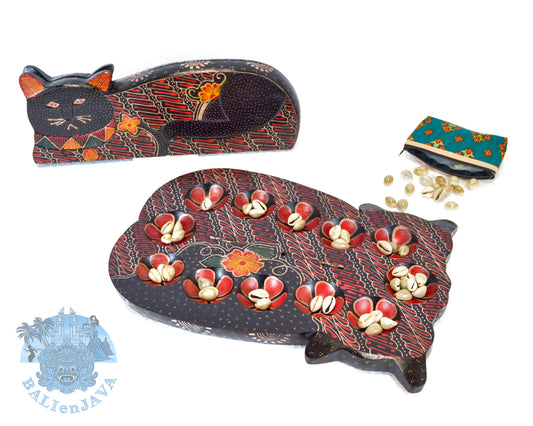 A wooden cat-shaped mancala board game set with red and yellow pattern, featuring two cat-shaped pieces, a teal coin purse, and cowrie shells scattered on a white background.