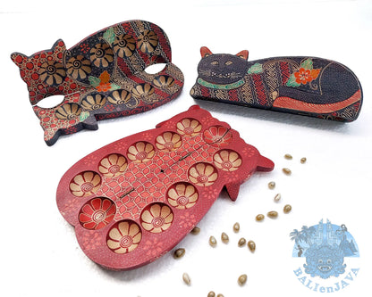 Three hand-painted wooden cat-shaped mancala game on a white background, including two closed board game with intricate floral patterns and one opened board game showing circular pits, with cowrie shells scattered nearby.