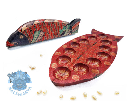 A wooden fish-shaped mancala board game set with red, green and black pattern, featuring two fish-shaped pieces, and cowrie shells scattered on a white background.