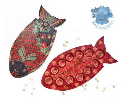 Two wooden fish-shaped mancala board game with red, green and black pattern and cowrie shells scattered on a white background.