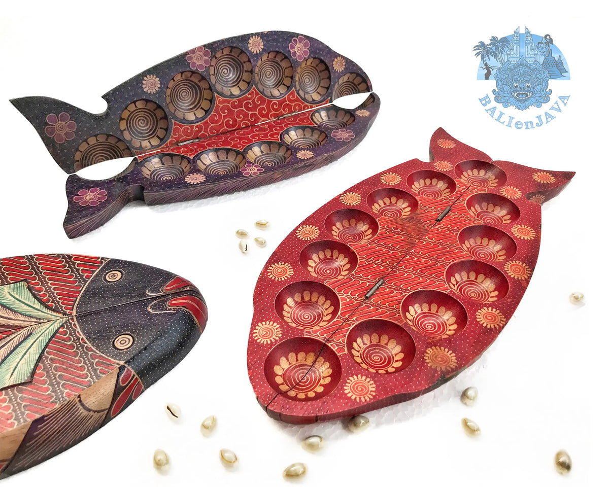 Three fish-shaped mancala board games with floral-patterned circular pits and cowrie shells scattered on a white background.
