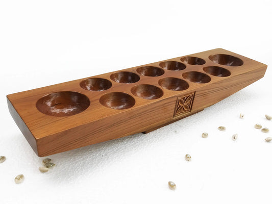 A wooden mancala board game with ten circular holes, photographed on a white surface with a few cowrie shells scattered nearby.