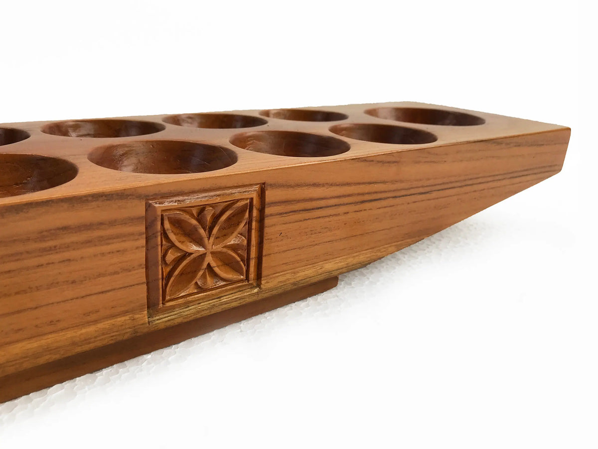 A close-up of a carved wooden mancala game board, showing a warm medium-brown wood grain, circular holes, and a decorative carved floral pattern on a white background.