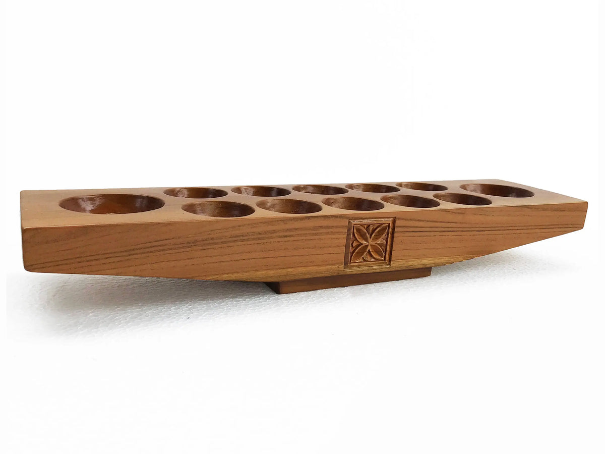 A carved wooden mancala game board, showing a warm medium-brown wood grain, circular holes, and a decorative carved floral pattern on a white background.