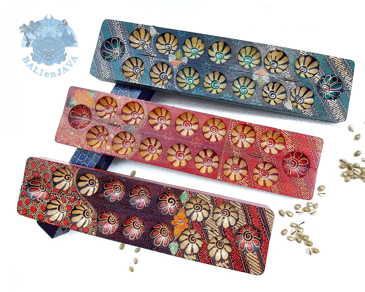 Three mancala board games with hand-painted design and seven circular pits in green, red, and brownish-red color, and cowrie shells scattered on a white background