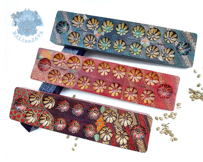 Three mancala board games with hand-painted design and seven circular pits in green, red, and brownish-red color, and cowrie shells scattered on a white background