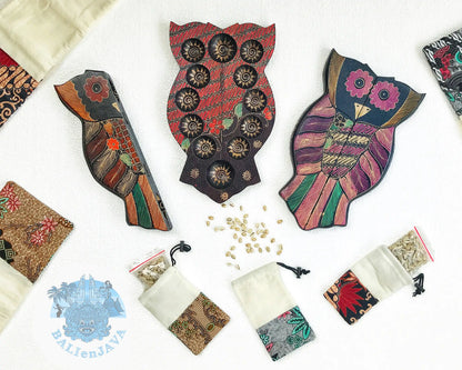 Three owl-shaped wooden mancala boards with various colors and patterns, surrounded by fabric pouches and cowrie shells scattered on white background.