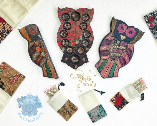 Three owl-shaped wooden mancala boards with various colors and patterns, surrounded by fabric pouches and cowrie shells scattered on white background.