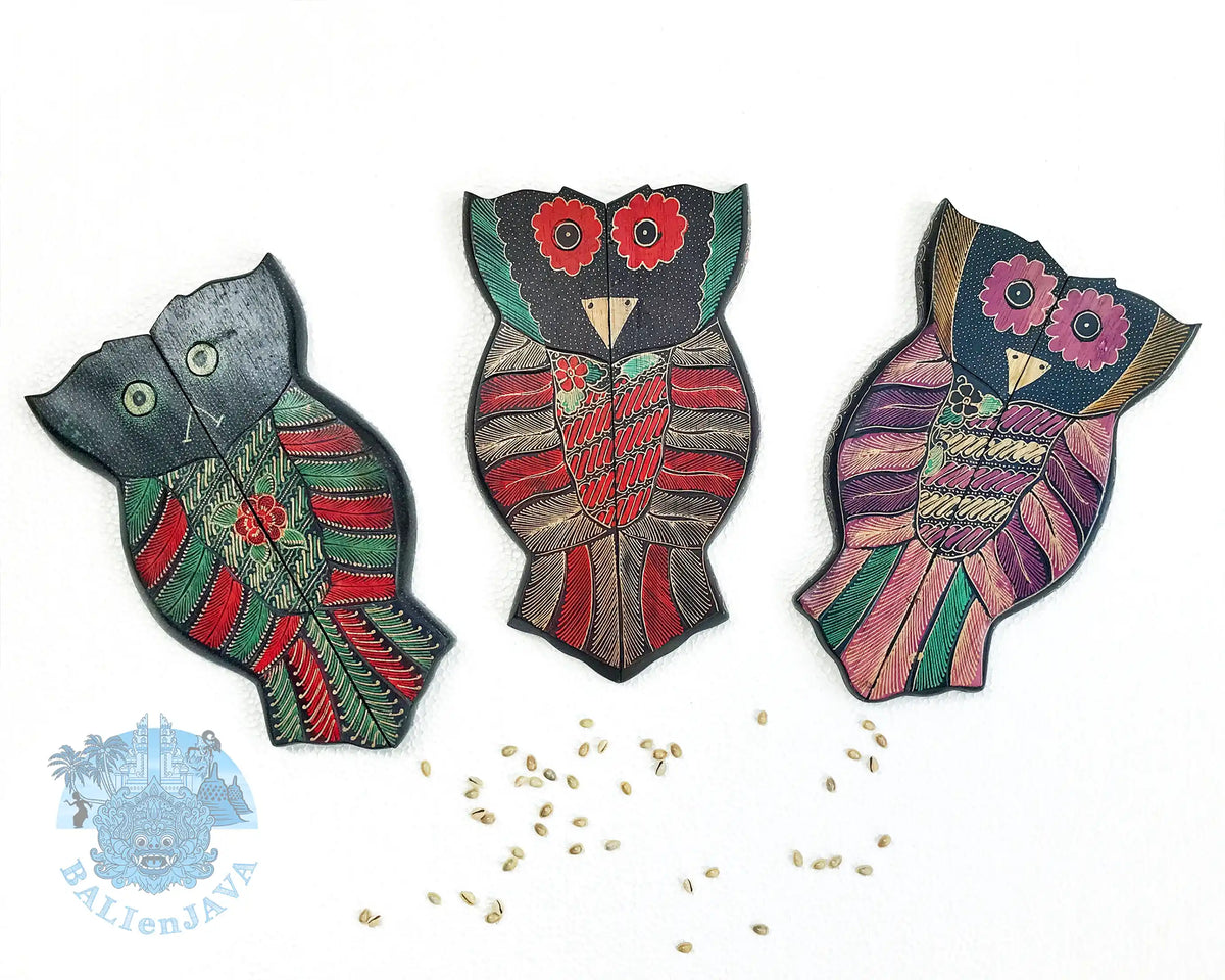 Three owl-shaped wooden mancala boards with various colors and patterns, with cowrie shells scattered on a white background.