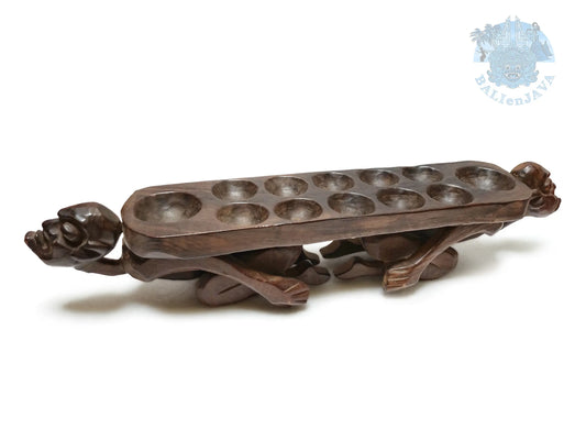 A dark brown carved wooden game board featuring human figures at each end and multiple holes for playing, photographed on a plain white background.