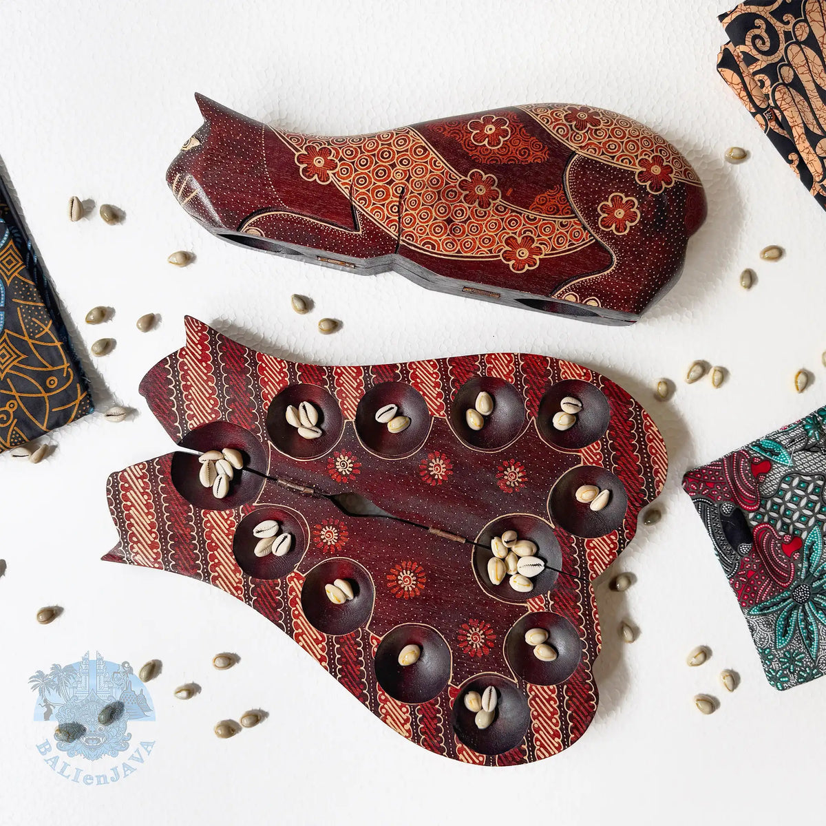 Two cat-shaped board games with brown hand-painted patterns, displayed on a white background with scattered cowrie shells. One board game is closed and the other is open, showing multiple small  circular pits holding cowrie shells inside.