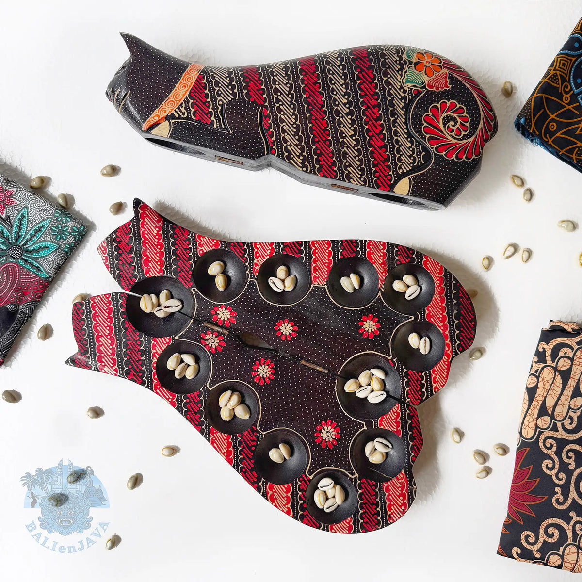 Two cat-shaped board games with intricate red, black, and beige batik patterns, arranged on a white surface with scattered cowrie shells and partially-visible fabric. 