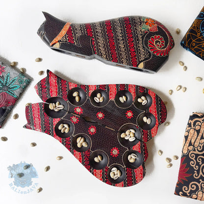 Two cat-shaped board games with intricate red, black, and beige batik patterns, arranged on a white surface with scattered cowrie shells and partially-visible fabric. 