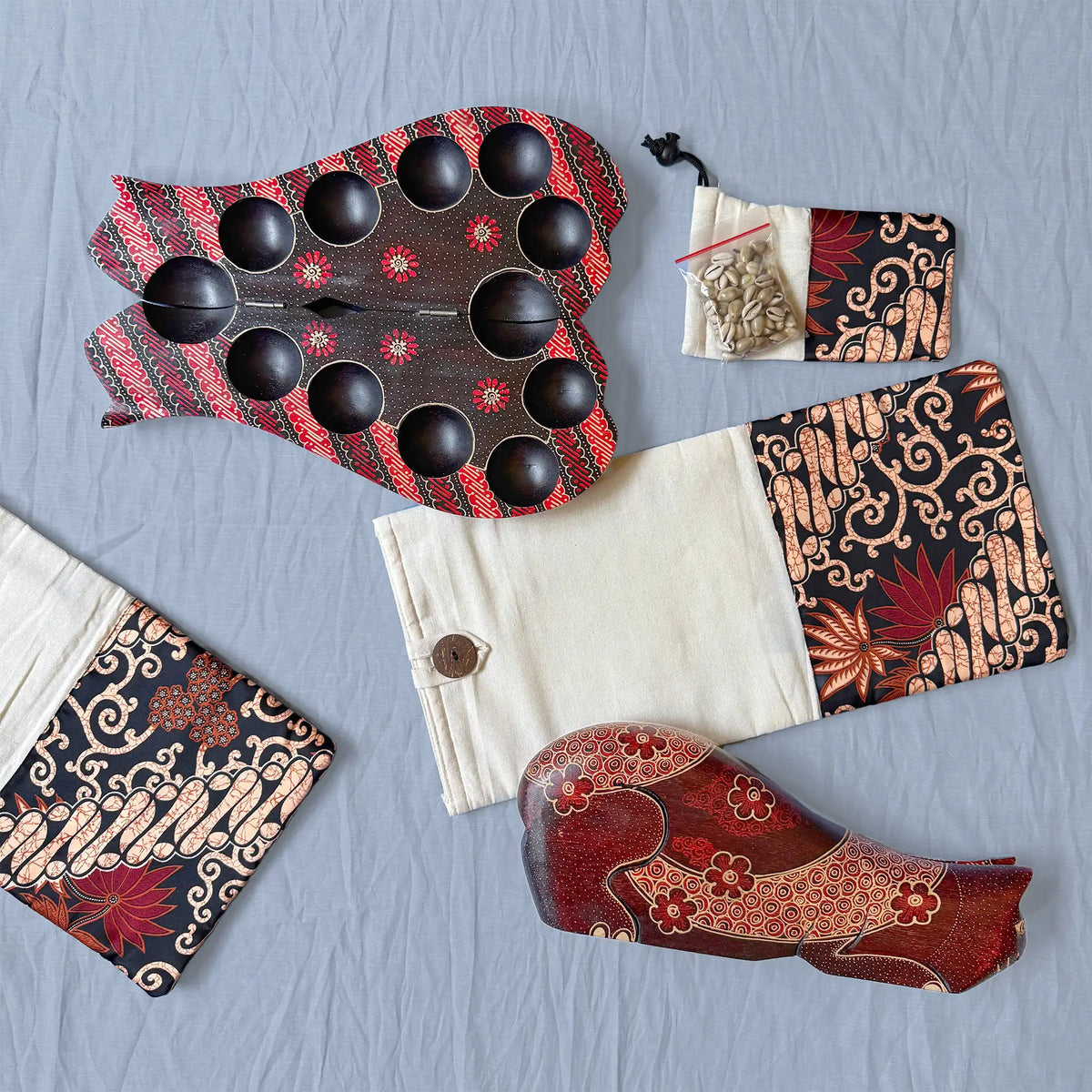 A set of two wooden board games with circular pits, a small pouch of cowrie shells, and patterned fabric pouch, arranged on a light blue textured background.