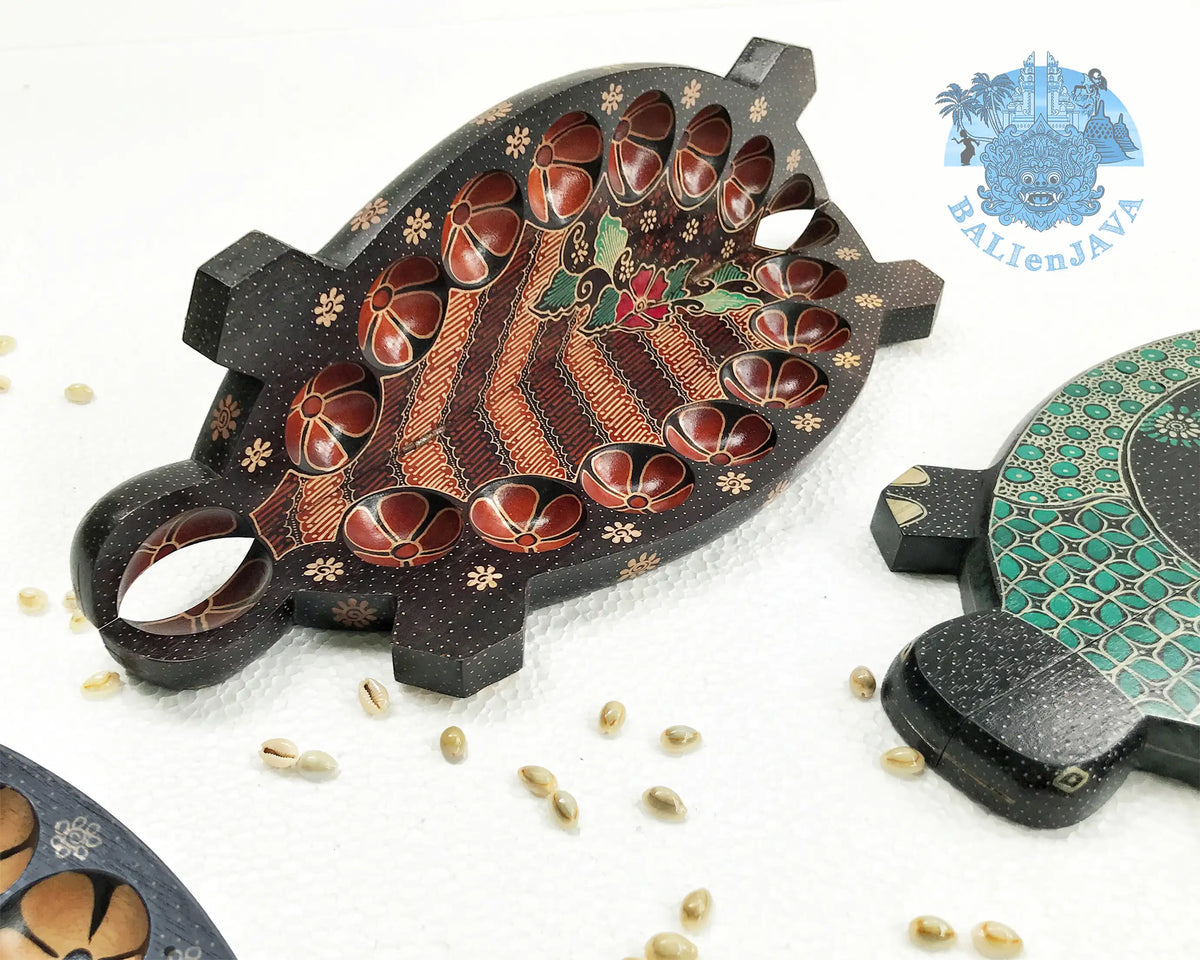 A brown turtle-shaped mancala board game on a white background in half-opened position, showing the circular pits on the inside, a green turtle board game is partially visible on the right side, with cowrie shells scattered nearby.