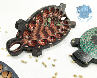 A brown turtle-shaped mancala board game on a white background in half-opened position, showing the circular pits on the inside, a green turtle board game is partially visible on the right side, with cowrie shells scattered nearby.