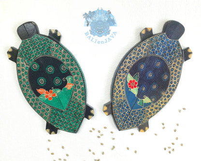 Two hand-painted turtle-shaped wooden mancala game in green and blue color on a white background, with cowrie shells scattered nearby.