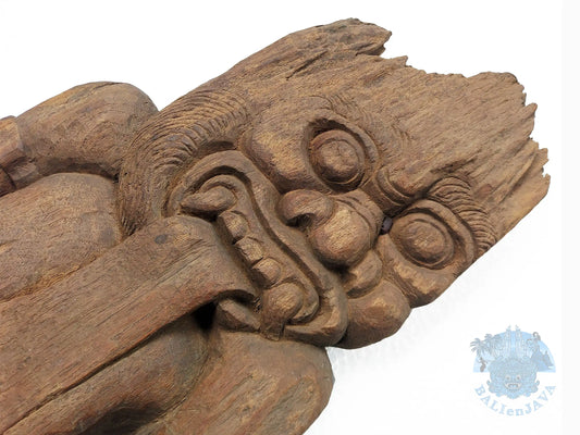 Close-up of a carved wooden sculpture showing a mythological figure with large circular eyes, textured wood grain, and a white background. 