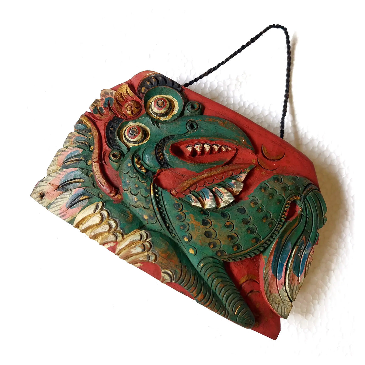 A hand-carved wooden bell depicting a mythological dragon with an open mouth, painted in green, red, and golden tones, suspended by a black cord against a plain white background.