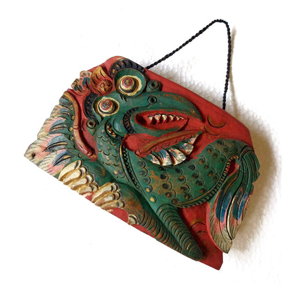 A hand-carved wooden bell depicting a mythological dragon with an open mouth, painted in green, red, and golden tones, suspended by a black cord against a plain white background.