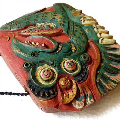 A close-up of a hand-carved wooden bell depicting a stylized dragon head, showcasing intricate hand-painted details in green, red, gold, and beige on a white background.