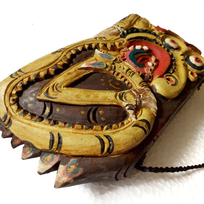 A carved wooden bell with a gold-painted outer frame, dark brown inner layers, and red, blue, and black painted details, shown on a white textured background.