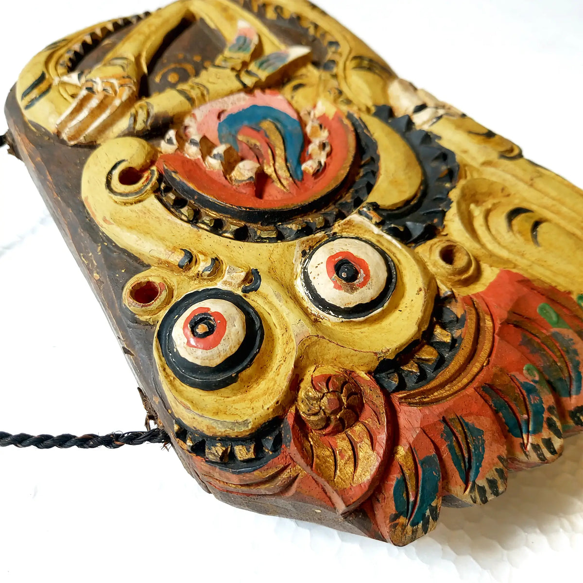 A close-up of a hand-carved wooden bell with an owl-like face, painted in gold, red, black, and teal on a white background.