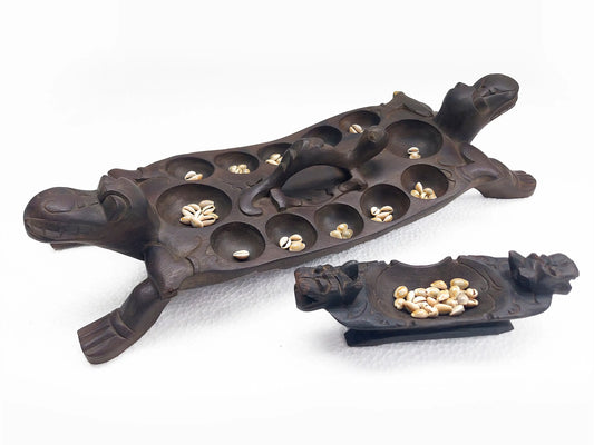 A dark carved wooden game board shaped like a dragon, with multiple holes containing cowrie shells, shown alongside a smaller matching plate on a white background.
