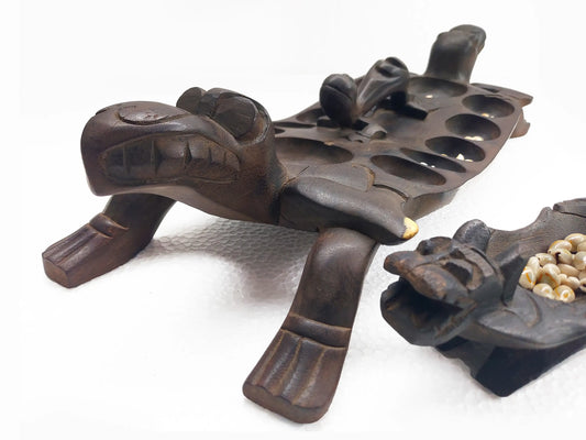 A carved dark wood game board and small plate set on a white background, featuring a main board with circular holes and a smaller matching plate showing cowrie shells.