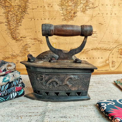 A vintage charcoal iron with a carved oriental dragon panel and wooden handle, displayed on a textured fabric surface in front of a vintage world map background.