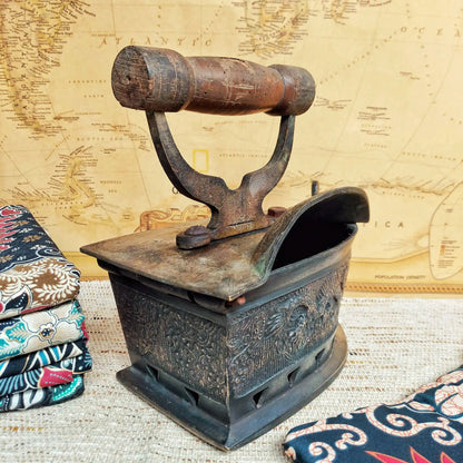 A vintage charcoal clothes iron with a dark brown wooden handle, displayed on a textured beige surface with a vintage map background and patterned fabric props.