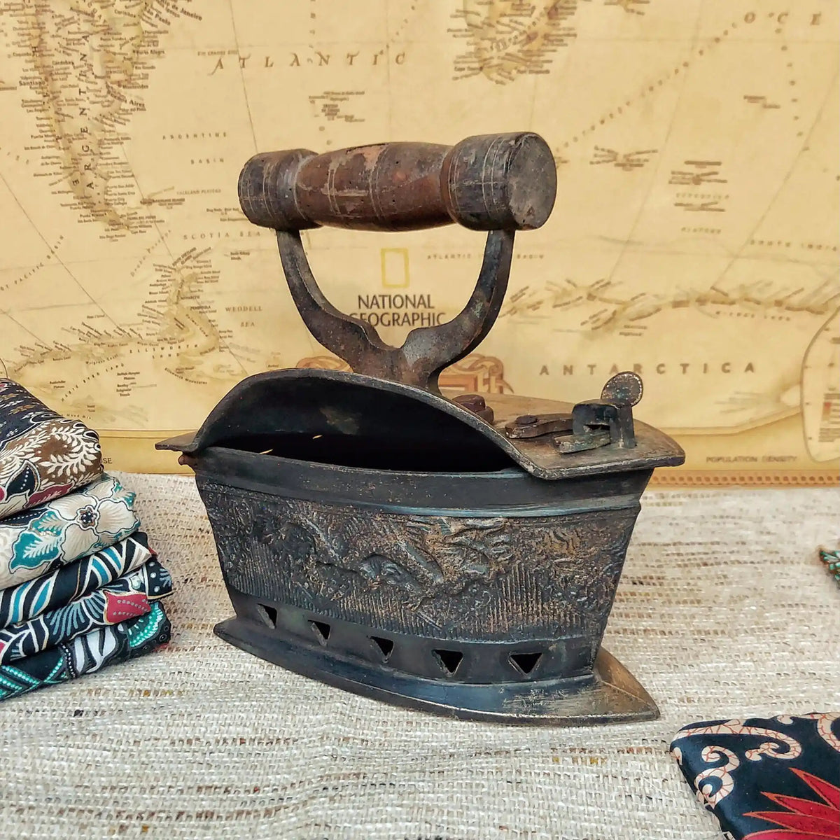 A vintage charcoal iron with dragon carving and a weathered wooden handle, displayed on a textured beige fabric surface in front of a vintage National Geographic map.