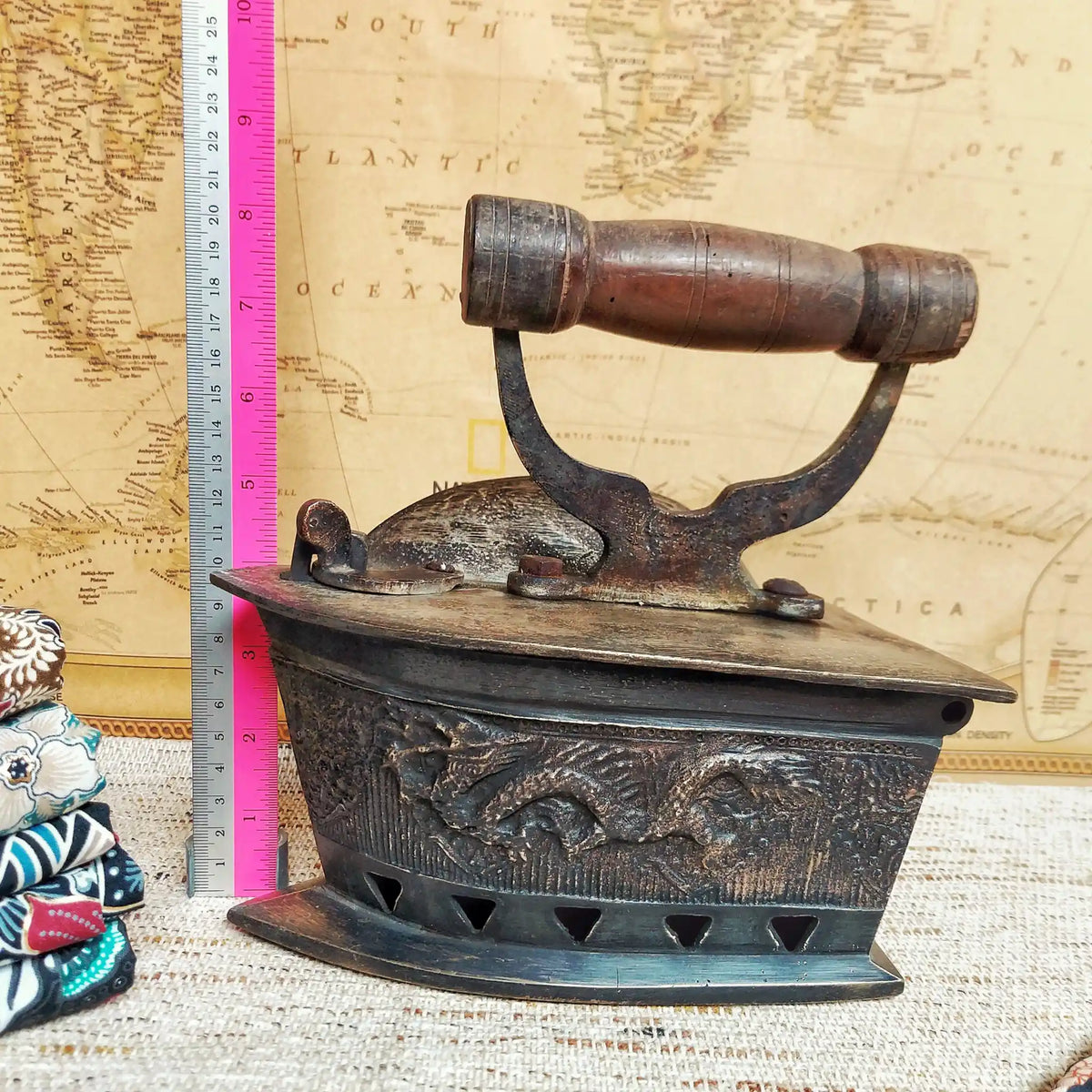 A vintage charcoal iron with a dark brown wooden handle, displayed next to a pink and silver ruler for scale against a vintage map background.